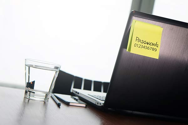 An office desk with a laptop that has an access password stuck to the back on a sticky note.